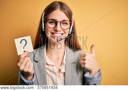 Young redhead call center agent woman using headset holding question mark reminder happy with big smile doing ok sign, thumb up with fingers, excellent sign