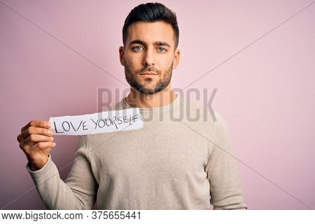 Young handsome man holding paper with self love message over pink background with a confident expression on smart face thinking serious