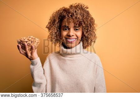 Young african american woman with curly hair holding bowl with healthy peanuts with a happy face standing and smiling with a confident smile showing teeth