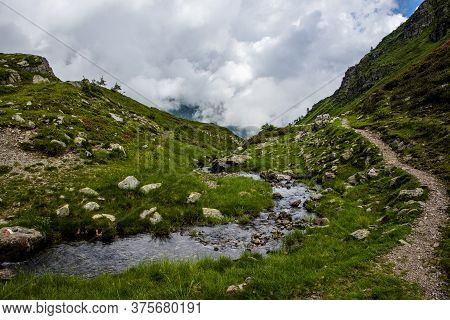 Stream Flows Placidly Among The Green Meadows On The Alpine And Granite Rocks Of The Lagorai Near La