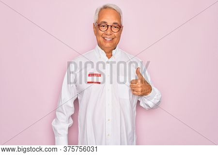 Middle age senior grey-haired man wearing hello my name is introduction sticker happy with big smile doing ok sign, thumb up with fingers, excellent sign