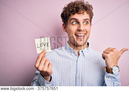 Young blond romantic man with curly hair holdiing paper with love message for valentines day pointing and showing with thumb up to the side with happy face smiling