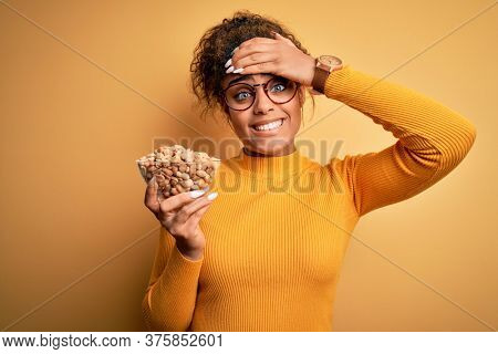 Young african american afro girl holding bowl with healthy peanuts over yellow background stressed with hand on head, shocked with shame and surprise face, angry and frustrated. Fear and upset