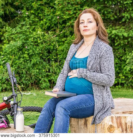 Pregnant Woman With Book Sitting In Green Park Outdoors Spting Time.