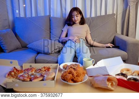Young Asian Woman Takeaway Eating Full Junk Food Sitting On Sofa And Watch Television In Living Room