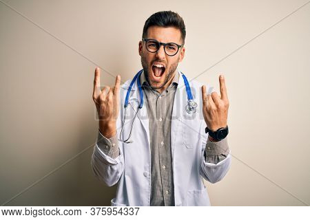 Young doctor man wearing glasses, medical white robe and stethoscope over isolated background shouting with crazy expression doing rock symbol with hands up. Music star. Heavy concept.