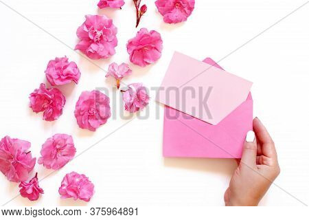 Hand Keeps Pink Envelope Near Pink Flowers  Over A White Background Top View