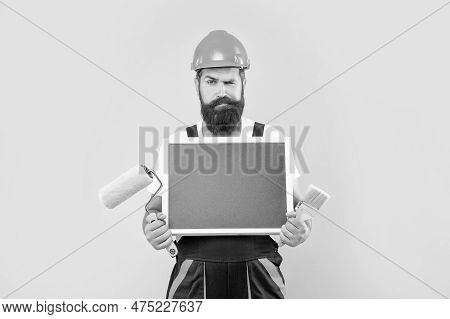 Man Housepainter In Helmet And Work Clothes Hold Paint Roller Brush And Blackboard With Copy Space