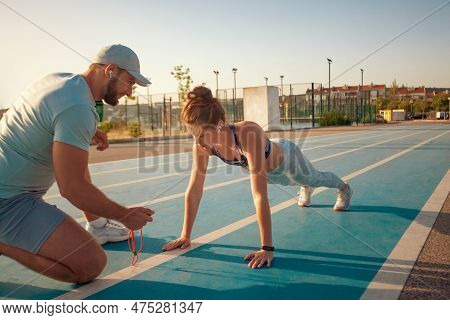 Instructor And Athlete Runner On The Track. The Athletics Marks The Time In The Plank Exercise On A 