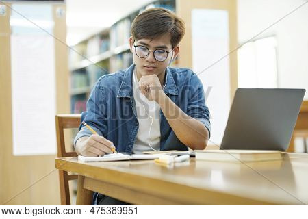 Young Male College Student Wearing Eyeglasses And In Casual Cloths Sitting At Desk Studying, Reading