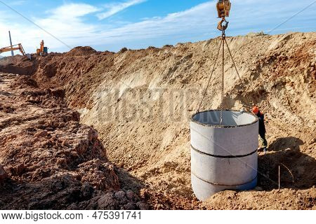 A Loader Lowers A Concrete Ring Into A Dug Hole To Build A Septic Tank. A Worker Installs A Sewer In