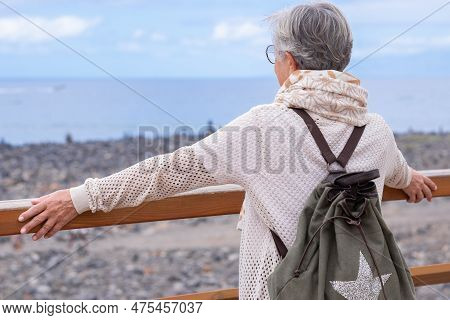 Rear View Of Senior Gray Haired Woman With Backpack Looking At Horizon Over Sea Enjoying Retirement,