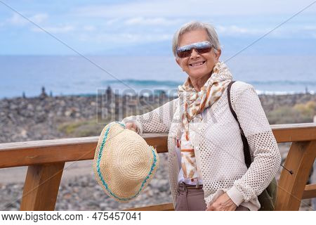 Happy Senior Gray Haired Woman With Backpack Walking Along The Sea Beach With Hat In Hand Enjoying V