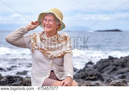 Beautiful Senior Gray Haired Woman With Hat Sitting On The Rock Beach At Sea Enjoying Vacation, Free