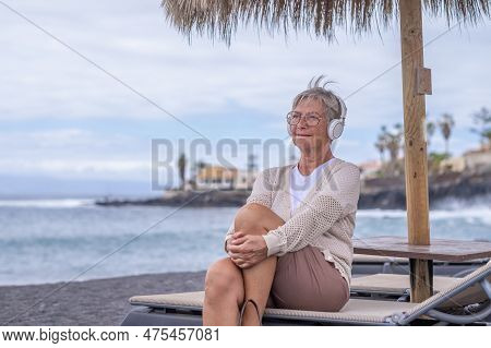 Smiling Caucasian Senior Woman With Headphones Sitting At The Beach Looking At The Horizon While Enj