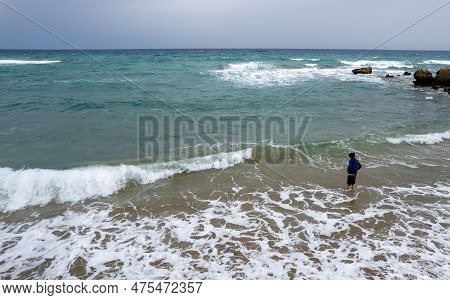 Drone Aerial Of Young Boy In Shorts Standing On The Summer Beach And Looking At The Stormy Spindrift