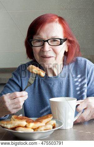 Grandmother Eating Pancakes And Drinking Tea Sitting At Kitchen Table In House. Elderly Woman With R