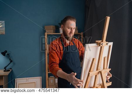 Portrait Caucasian Bearded Male Artist In An Apron In A Home Studio Stands Near An Easel Holds A Can