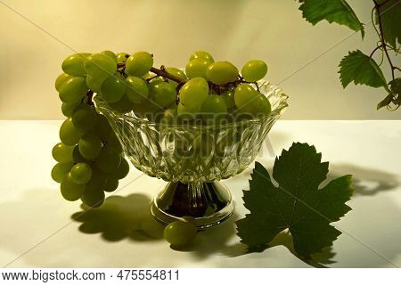 A Bunch Of White Grapes In A Glass Bowl With A Silver Base. A Grape Vine And Leaf Serve As Decoratio