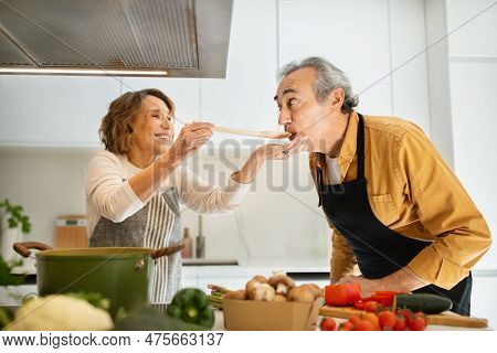 Happy Senior Spouses Tasting Food, Woman Giving Her Husband To Try Soup, Cooking Lunch Together In K