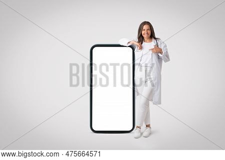 Positive Happy Senior Doctor Woman In White Uniform Posing And Pointing At Big Cellphone With White 