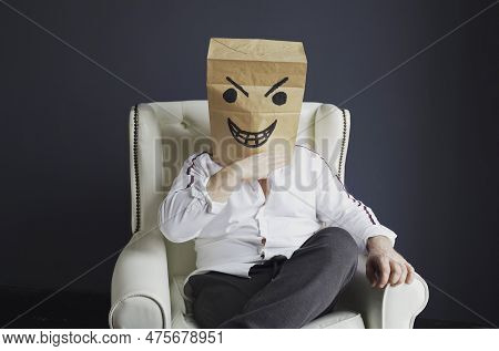 A Man With A Paper Bag On His Head, With An Evil Smiley Face Drawn, Sits On A White Chair, Gloats Em