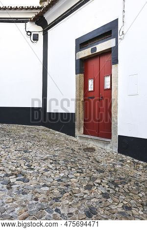 Typical Portuguese Facade With Red Wooden Door On Cobblestone Street In Evora Town, Portugal