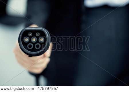 Focus Closeup Hand Holding Ev Plug For Electric Vehicle Pointing At Camera With Charging Station At 