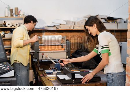 Cheerful Typographer Using Paper Trimmer Near Colleague Next To Professional Print Plotter.