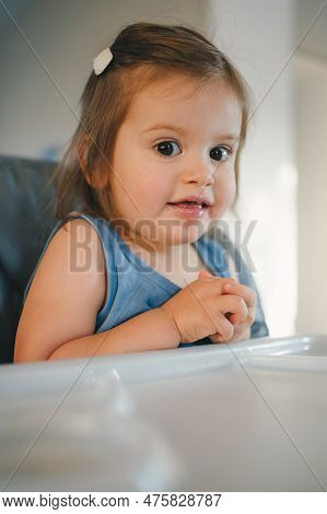 Close-up View Of A Child Sitting In A High Chair For Feeding In The Kitchen, Looking At C Amera And 