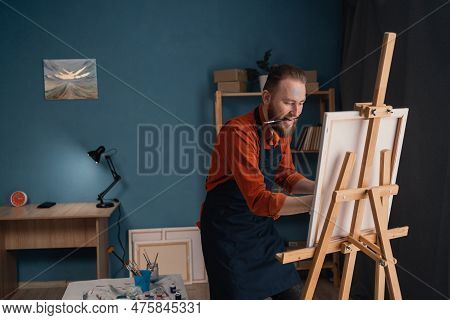Portrait Caucasian Bearded Male Artist In An Apron In A Home Studio Stands Near An Easel Holds A Can