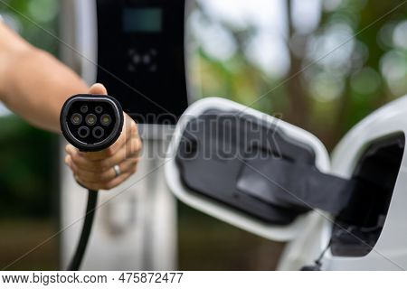 Focus Closeup Hand Holding Ev Plug For Electric Vehicle Pointing At Camera With Charging Station At 
