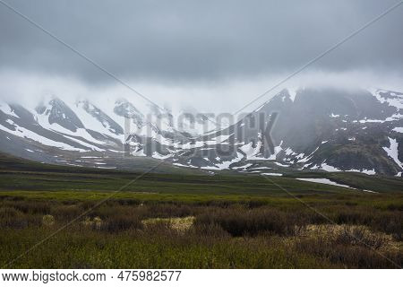 Dramatic Alpine Landscape With Snowy Mountains In Gray Low Clouds. Bleak Atmospheric Scenery Of Tund