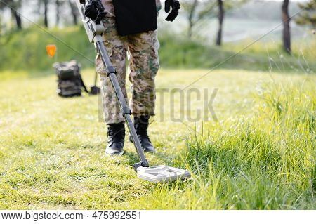 A Man In A Military Uniform And Bulletproof Vest Works In The Forest With A Metal Detector. A Minesw