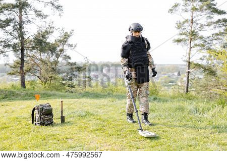 A Man In A Military Uniform And Bulletproof Vest Works In The Forest With A Metal Detector. A Minesw