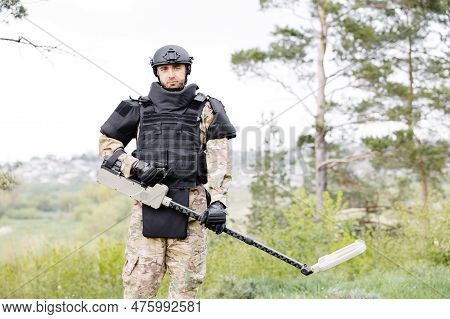 A Man In A Military Uniform And Bulletproof Vest Works In The Forest With A Metal Detector. A Minesw