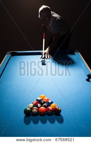Man playing a game of pool lining up as the break shooter as he takes aim with his cue and ball at the triangular formation of object balls in a dark shadowy pub or bar