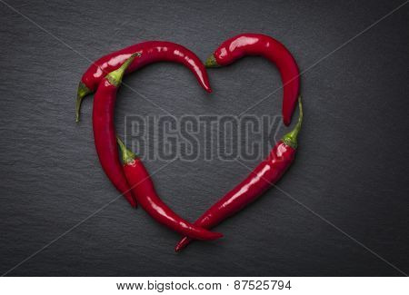 Red chili peppers in heart shape, indicating hot love for valentine's day, isolated on dark grey slate stone.