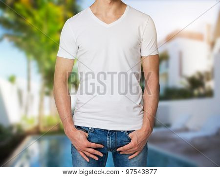 Young Man In A White V Shape T-shirt, Hands In Pockets. Villa With Swimming Pool In Blur On The Back
