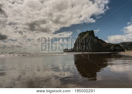 Auckland New Zealand - March 2 2017: Lion rock reflected in wet sandy Piha beach in Tasman Sea surf and under blue sky with white clouds. Some people.