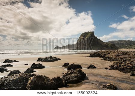 Auckland New Zealand - March 2 2017: Lion rock on Piha Beach of Tasman Sea surrounded by surf and under blue cloudy sky. Forefront is sand and dispersed black volcanic rocks.