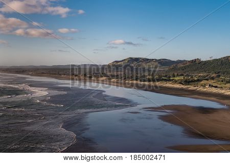 Auckland New Zealand - March 2 2017: Twilight wide shot photo of Muriwai Beach under blue sky reflected on wet beach invaded by Tasman Sea surf. Dunes mountains and more in back.