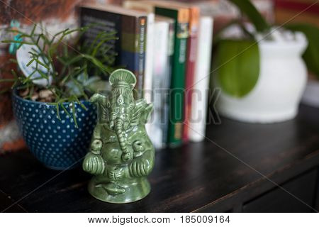 Home interior decor a ceramic statuette of Ganesh, books and flower pots with plants on black wooden commode on a red brick wall background