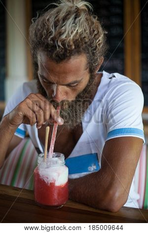Attractive bearded curly man with a mug of smoothie is sitting in a cafe drinking through a straw