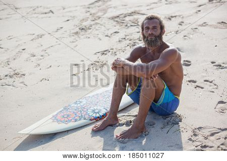 Attractive bearded curly man with a surfboard is seating on a sand and looking to the camera