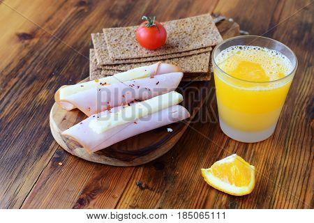 Wooden cutting board with crispbread, cheese, ham, cherry tomato, glass of fresh orange juice on a wooden background. Healthy breakfast. Healthy eating concept