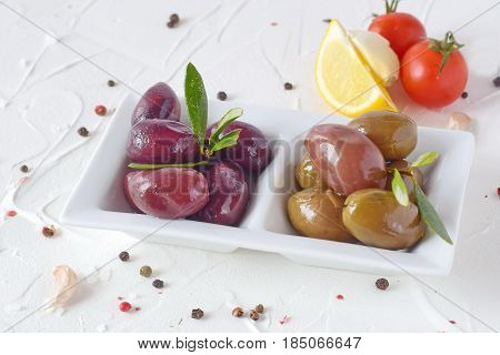 White plate with black and green olives with olive leaves on a white abstract background with lemon, cherry tomato, spices. Healthy food. Greek food. Mediterranean food