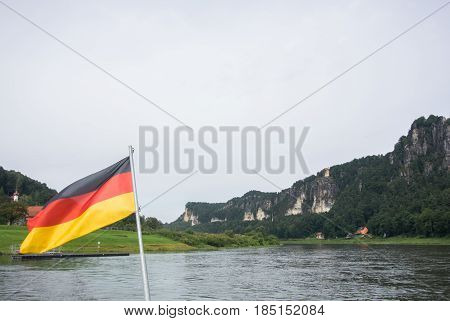 German Flag At Ferry Through The Elbe At Kurort Rathen Village And Bastei Rocks At The Background, S