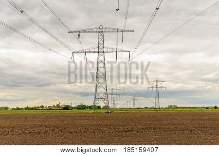 Rows of power pylons in a rural Dutch landscape. In the background is the power plant with a smoking chimney and a cooling tower. It is springtime now.