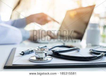 View Of Stethoscope And Equipment On Foreground Table With Doctor Using Computer Keyboard In Clinic